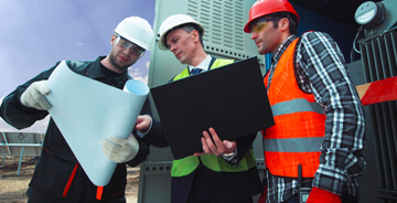 Three workers wearing hard hats and safety gear reviewing blueprints and a laptop at an industrial worksite.
